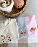 Fall sweaters showcasing cozy designs, including themes of autumn leaves, pumpkins, and the phrase 'In My Cozy Era'.